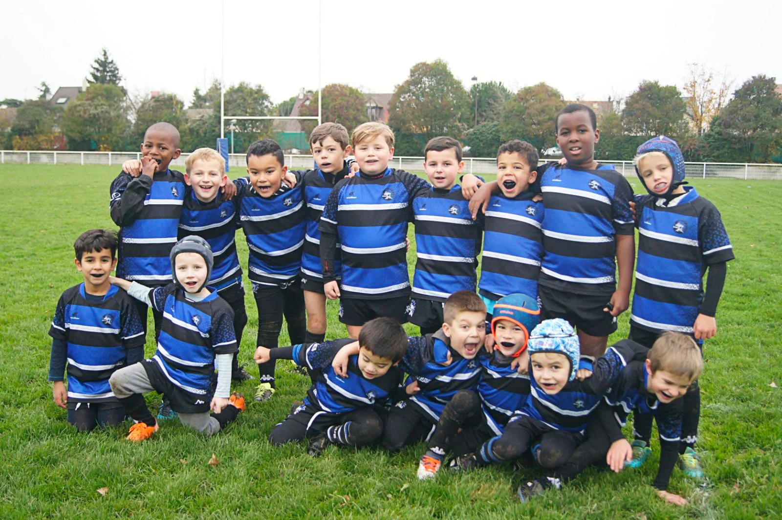 Un groupe de jeunes joueurs de l'école de rugby posent ensemble sur un terrain herbeux, vêtus de maillots rayés bleus et noirs. Ils se tiennent serrés les bras l’un autour de l’autre, souriant et riant, démontrant leur esprit d’équipe et leur camaraderie. Certains joueurs sont agenouillés devant, ajoutant à l'ambiance ludique et animée. Les expressions des enfants reflètent leur plaisir et leur enthousiasme pour le jeu, créant une scène réconfortante et joyeuse.