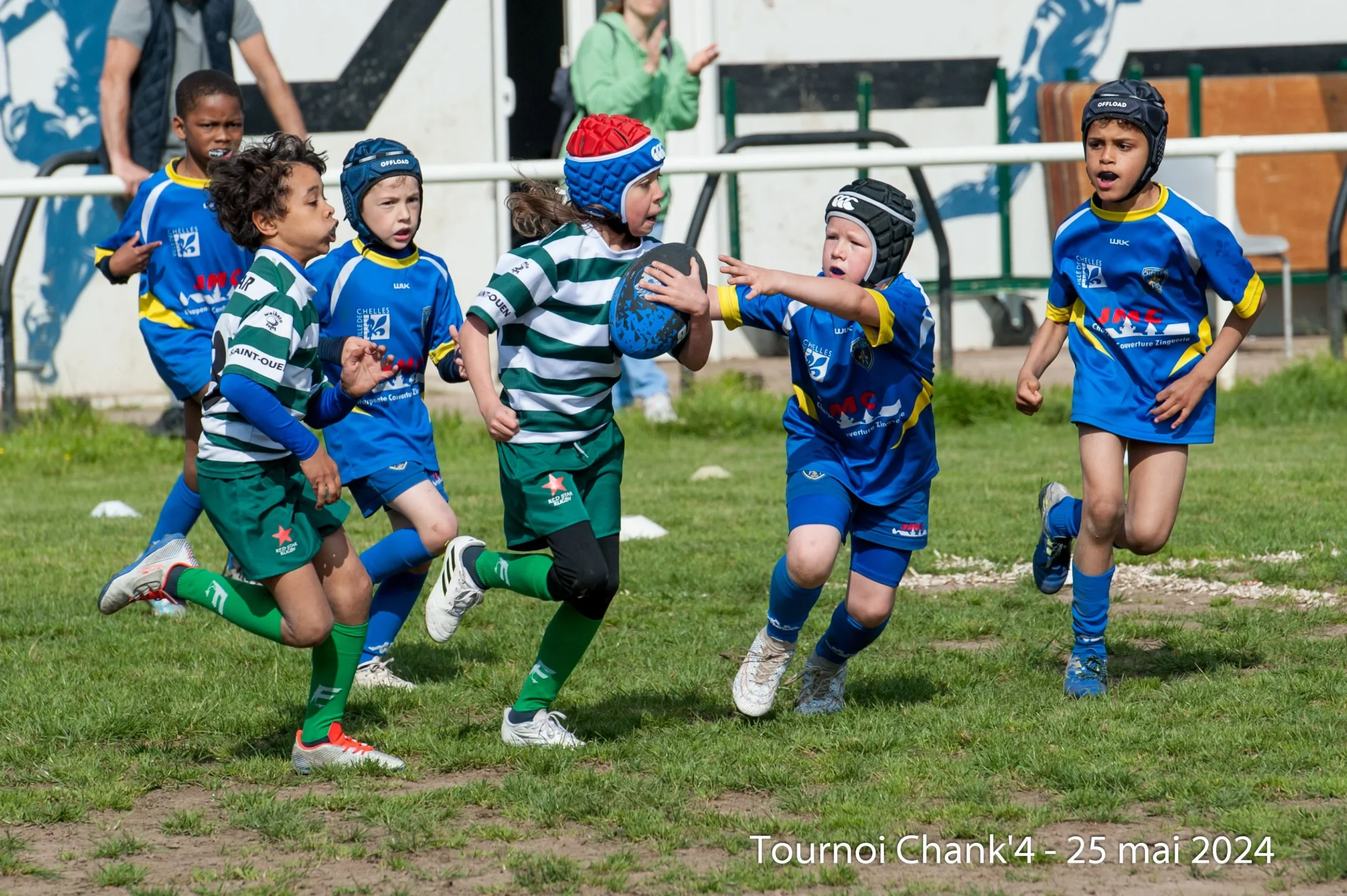 De jeunes rugbymen s'affrontent avec énergie lors d'un match lors du "Tournoi Chank'4", le 25 mai 2024. Un joueur en maillot rayé vert et blanc court avec le ballon, face à l'opposition déterminée des joueurs en maillot bleu et jaune. L'un des joueurs vêtus de bleu tente de tacler en tendant la main vers le porteur du ballon. Les jeunes athlètes portent un casque de protection et font preuve d’une concentration intense lorsqu’ils participent au jeu. La scène capture l’enthousiasme et la compétition animée du rugby des jeunes.