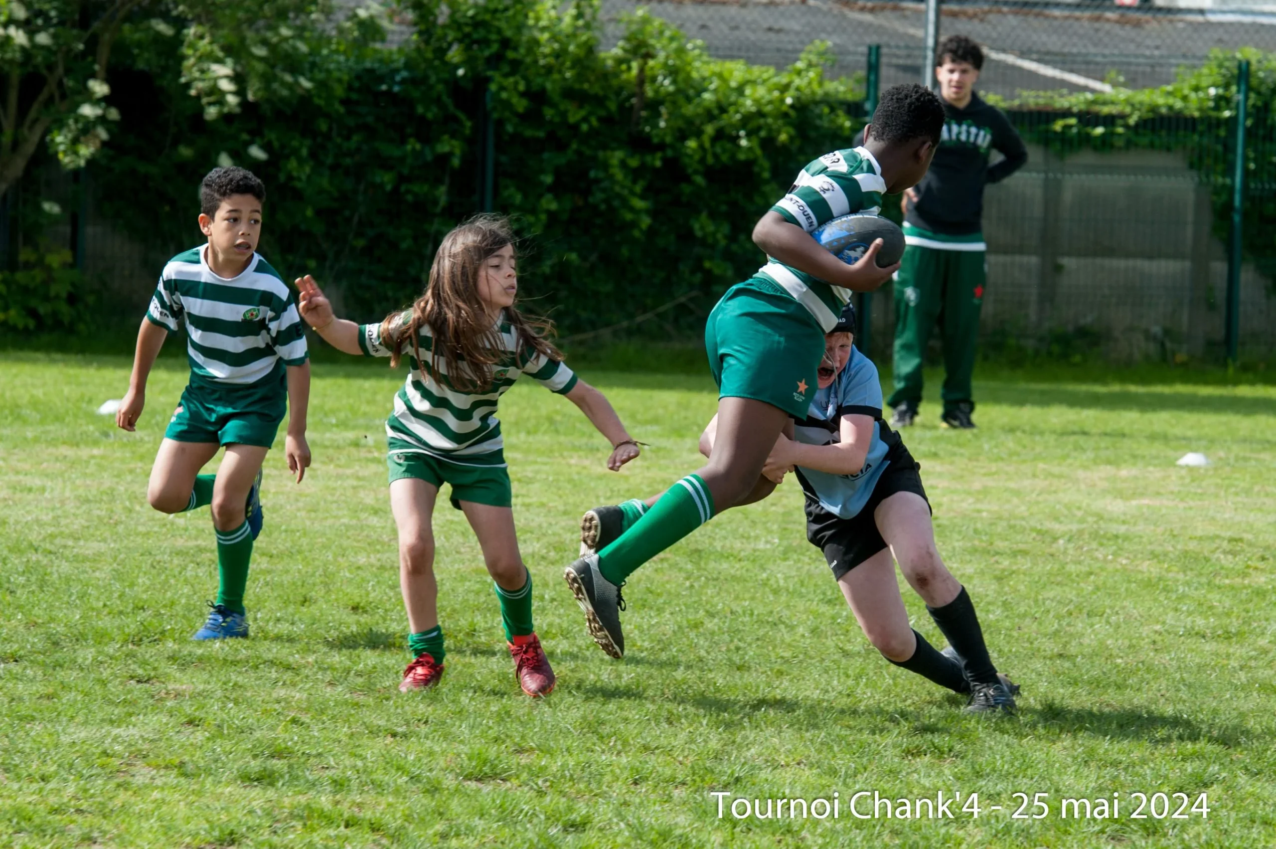 Lors du « Tournoi Chank'4 » le 25 mai 2024, de jeunes rugbymen sont profondément engagés dans un match. Un joueur vêtu d'un maillot rayé vert et blanc sprinte avec le ballon, tentant d'échapper à un adversaire déterminé d'une autre équipe qui tente de plaquer. Deux coéquipiers, également vêtus de maillots rayés verts et blancs, courent juste derrière, prêts à soutenir leur coéquipier. Les spectateurs et les entraîneurs observent l'action depuis les coulisses, capturant l'intensité et l'enthousiasme du rugby des jeunes.