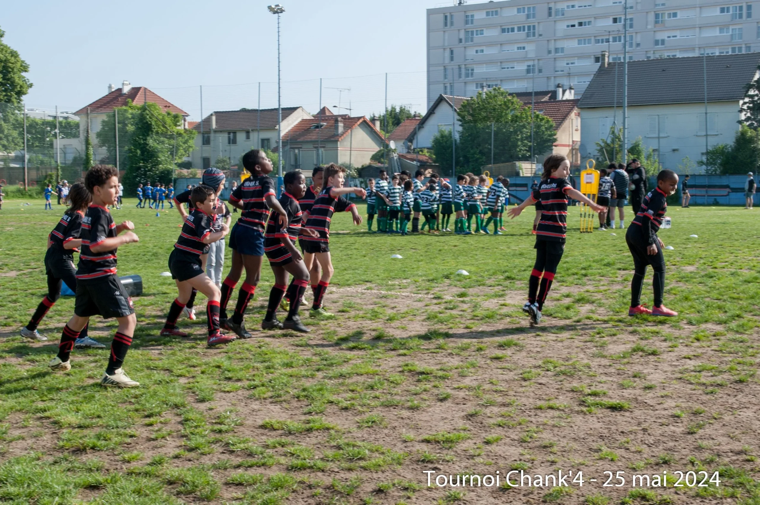 Lors du « Tournoi Chank'4 » le 25 mai 2024, de jeunes rugbymen participent à une séance d'échauffement. Une équipe vêtue de maillots rayés noirs et rouges s'entraîne sur le terrain, engagée dans des exercices dynamiques pour préparer son match. En arrière-plan, une autre équipe vêtue de maillots rayés verts et blancs se rassemble pour une discussion d'équipe, tandis que des joueurs vêtus de maillots bleus sont visibles plus loin, se préparant également pour leur match. La scène capture la nature énergique et organisée du tournoi, mettant en valeur l'enthousiasme et le dévouement des jeunes athlètes.