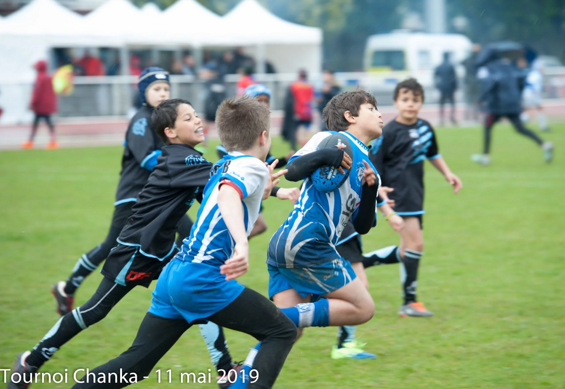 Un groupe de jeunes joueurs de rugby se trouve au milieu d'un match intense sur un terrain herbeux. Le joueur au premier plan, vêtu d'un maillot bleu et blanc, tient fermement le ballon de rugby et sprinte vers l'avant, tandis que des joueurs en maillot noir tentent de le plaquer. Les expressions sur leurs visages montrent la détermination et l'effort. La scène capture l'action, la compétition et l'excitation du rugby des jeunes.