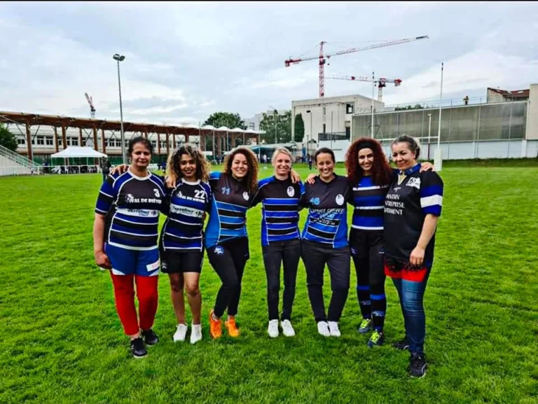 Un groupe de sept rugbywomen de Pôle compétition rugby se tient debout sur un terrain herbeux et pose pour une photo. Ils portent tous des maillots de rugby assortis en bleu et noir. Les femmes sourient et se tiennent dans les bras, faisant preuve de camaraderie et d’esprit d’équipe. La scène capture la joie et l'unité de l'équipe féminine de rugby.