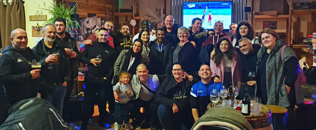 Un grand groupe de bénévoles est rassemblé dans un cadre intérieur confortable et pose pour une photo de groupe. L'atmosphère est chaleureuse et festive, avec des gens souriants, tenant un verre et faisant preuve d'un sentiment de camaraderie. En arrière-plan, un écran de télévision diffuse un événement sportif. L'espace présente un décor rustique avec des éléments en bois, des plantes et des décorations diverses. Le groupe comprend des hommes, des femmes et des enfants, tous habillés de façon décontractée, suggérant une réunion sociale ou une célébration décontractée. Des bouteilles et des verres sont posés sur une table au premier plan, renforçant l'ambiance conviviale.