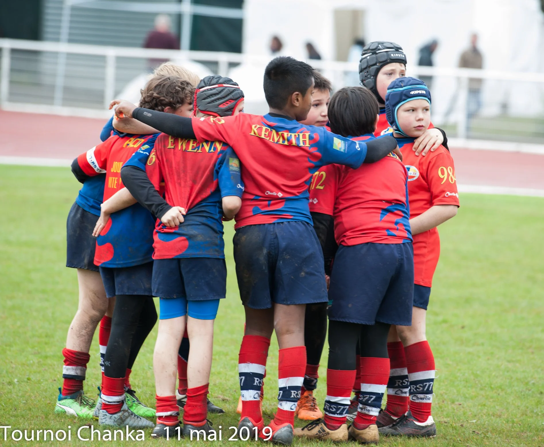 Un groupe de jeunes joueurs de rugby se rassemblent sur le terrain, les bras enroulés les uns autour des autres dans une démonstration d'unité d'équipe et de camaraderie. Les enfants portent des maillots rouges et bleus assortis avec différents numéros et noms au dos, ainsi qu'un short noir et des chaussettes rouges. Trois joueurs portent un casque de protection. La scène capture les liens étroits et le travail d’équipe entre les jeunes athlètes.
