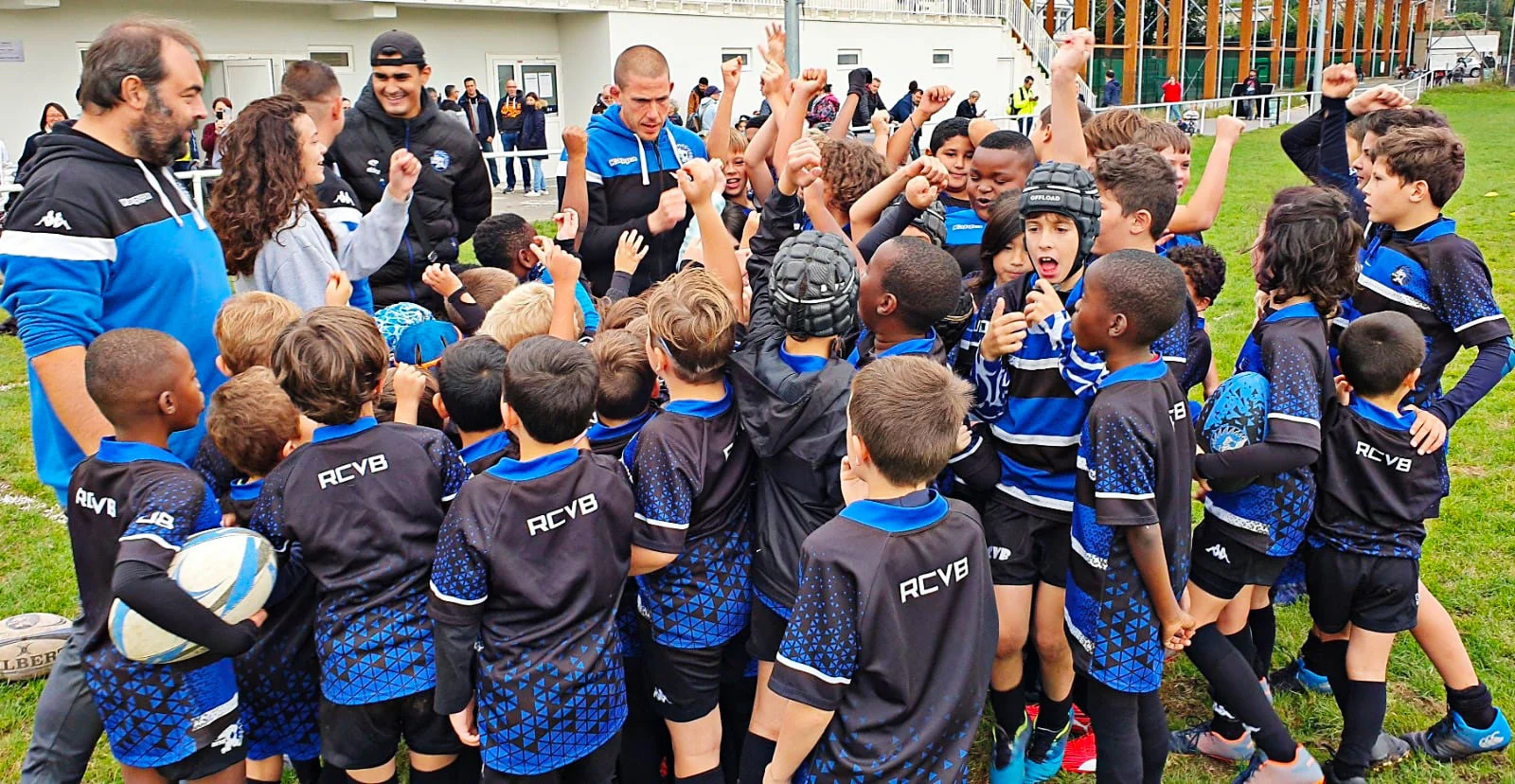 Jeunes joueurs de notre rugby club et leurs entra&icirc;neurs se rassemblent, levant la main. Les enfants portent des maillots noirs et bleus avec &laquo; RCVB &raquo; imprim&eacute; dans le dos, certains tiennent des ballons de rugby et portent des casques de protection. Les entra&icirc;neurs, v&ecirc;tus des tenues de leur &eacute;quipe, interagissent avec enthousiasme avec les enfants et m&egrave;nent les acclamations. La sc&egrave;ne capture un moment d&rsquo;esprit d&rsquo;&eacute;quipe et d&rsquo;enthousiasme, mettant en valeur l&rsquo;&eacute;nergie et la camaraderie des jeunes athl&egrave;tes et de leurs mentors.