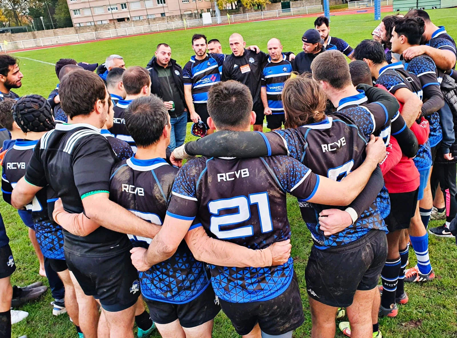 L'&eacute;quipe de P&ocirc;le comp&eacute;tition rugby RCVB est rassembl&eacute;e en groupe sur un terrain herbeux, avec des joueurs v&ecirc;tus d'uniformes noirs et bleus. Les joueurs ont les bras autour des &eacute;paules, d&eacute;montrant leur unit&eacute; et leur travail d'&eacute;quipe. Plusieurs entra&icirc;neurs et membres de l'&eacute;quipe se tiennent au milieu de la r&eacute;union, donnant un discours d'encouragement ou une discussion strat&eacute;gique. L'atmosph&egrave;re est concentr&eacute;e et &eacute;nergique, typique d'une r&eacute;union d'&eacute;quipe d'avant-match ou de mi-temps.